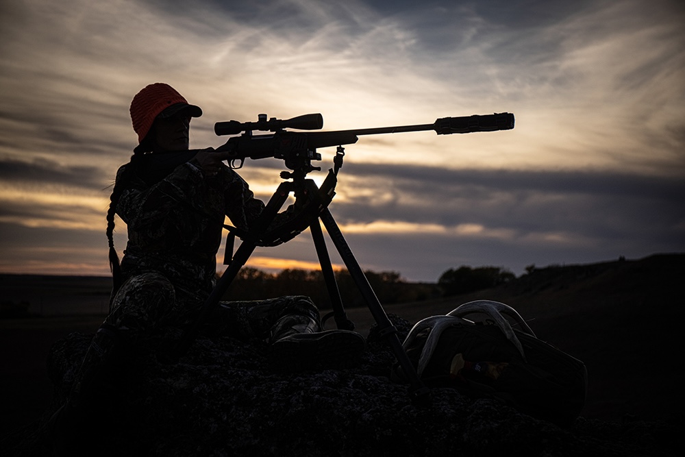 Silhouette of a hunter with a suppressed rifle on a tripod in the field.